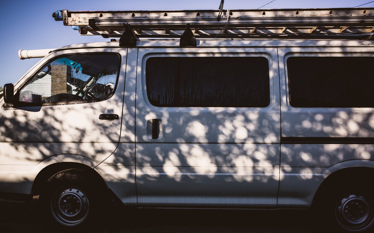 A white van with a ladder on top casting intricate shadows from nearby trees outdoors.